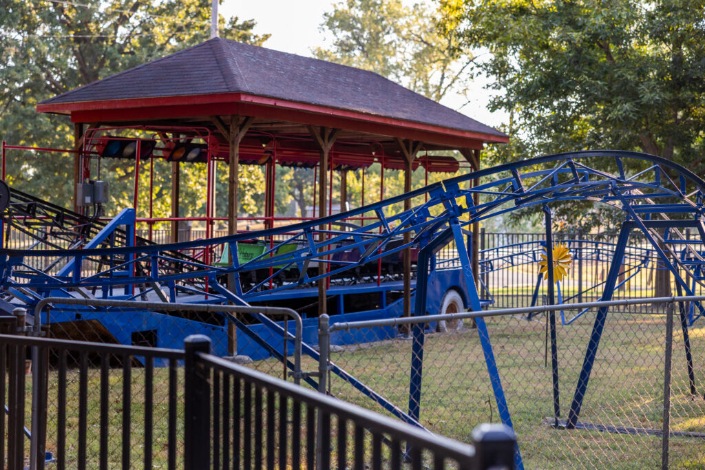 A freshly repainted blue kiddie roller coaster at Pittsburg's Kiddie Land, set behind a black metal fence with a covered pavilion in the background